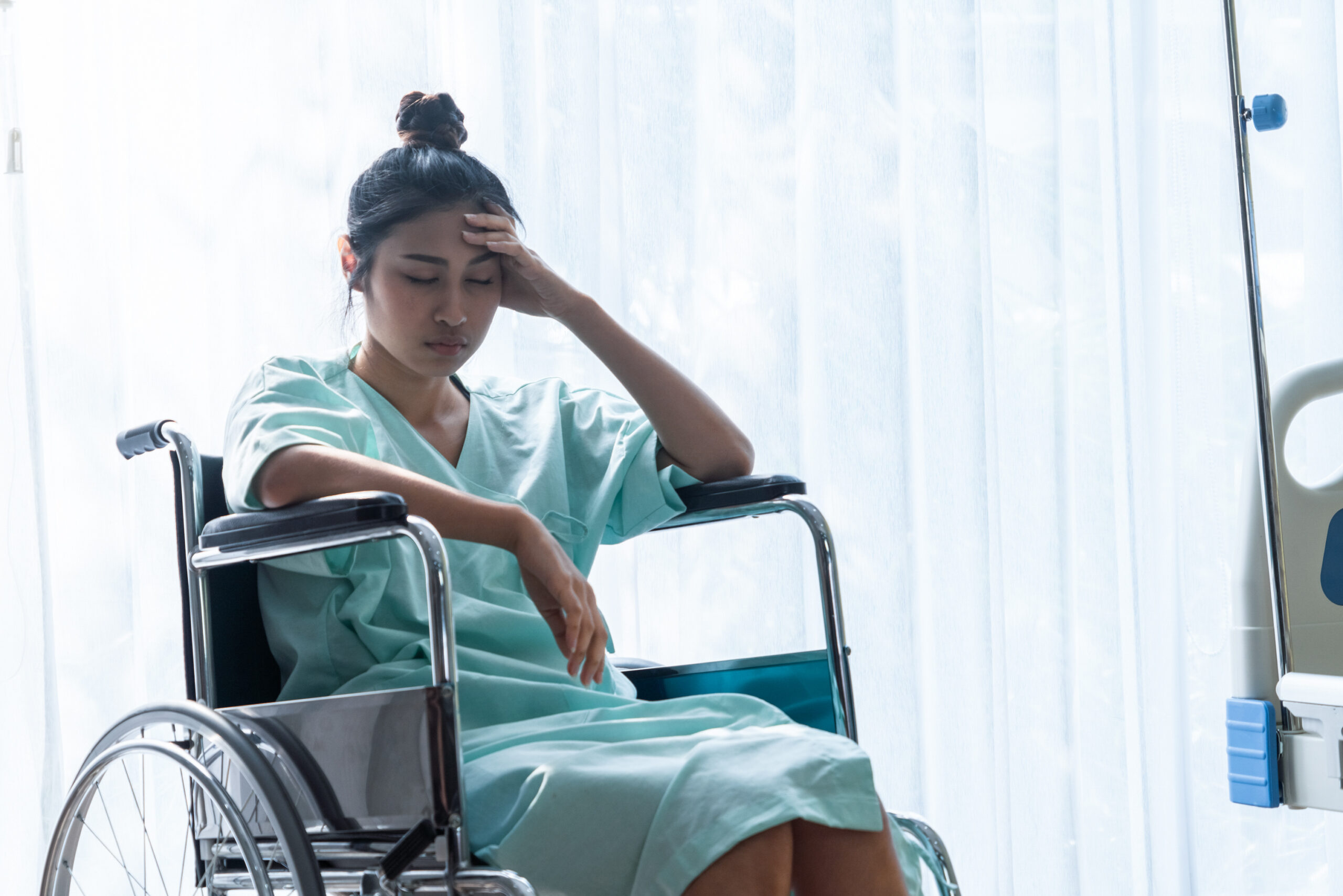 woman sitting in a wheelchair with her hand on her head and her eyes closed, showing signs of dizziness and fatigue. 