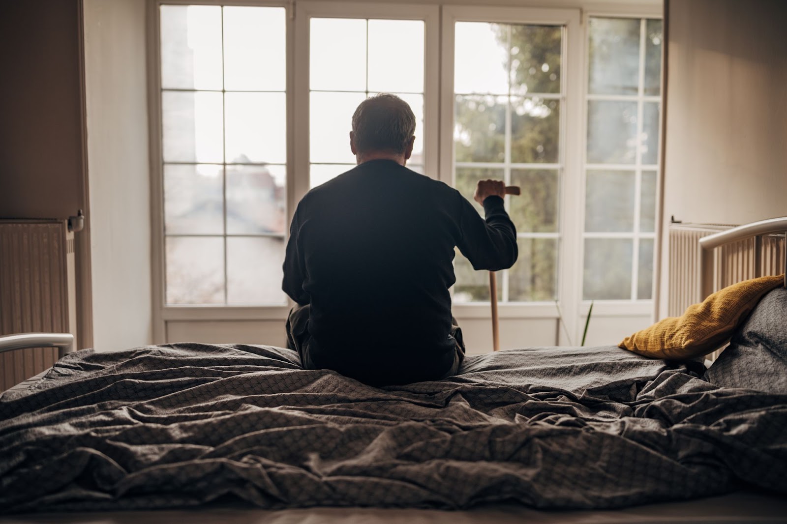 An older man sits on the edge of his bed, holding his cane and looking out the window. 