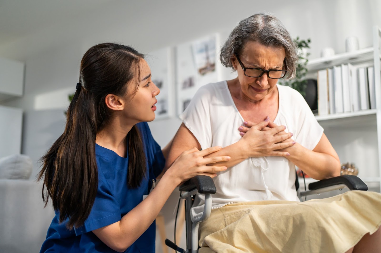 An aide offers assistance to an older woman in a wheelchair who is experiencing discomfort and is holding her hands over her heart.