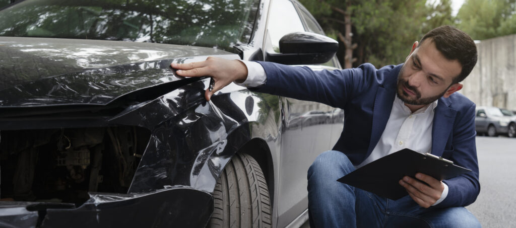 An insurance adjuster reviews the damage to the front end of a black car after a car accident. 